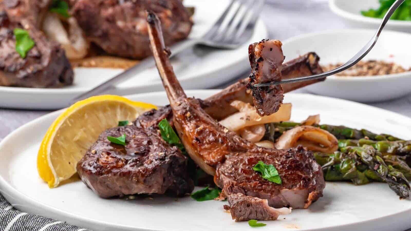 Plate of grilled lamb chops with asparagus, onions, parsley garnish, and a lemon wedge. A fork holds a piece of lamb.