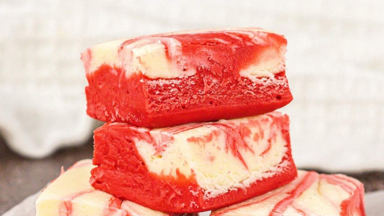 Three rectangular pieces of red and white swirled fudge are stacked on top of each other, with a soft, creamy texture visible. The background is neutral and out of focus.