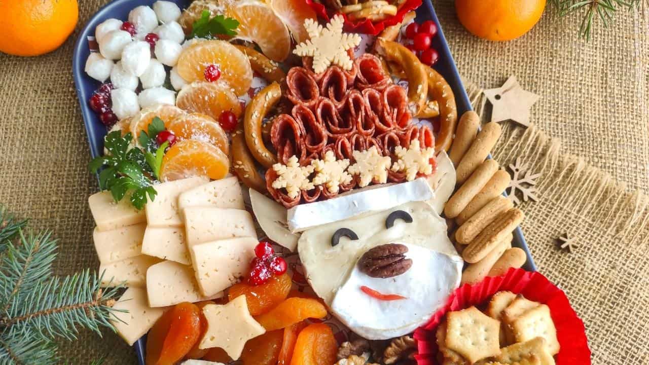A festive charcuterie board features cheese, crackers, dried fruit, pretzels, and orange slices, arranged with brie cheese shaped like a bear’s face, decorated with nuts and herbs. Oranges and pine branches are nearby.