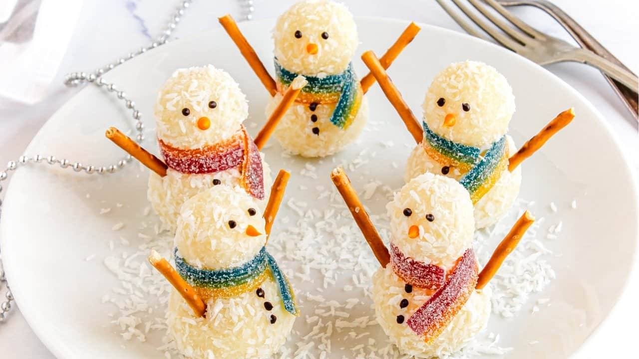 Five snowmen-shaped treats made of white coconut balls, decorated with pretzel stick arms, candy scarves, small candy facial features, and buttons, arranged on a white plate with scattered coconut flakes. Forks are in the background.