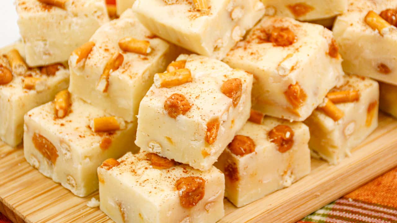 A collection of creamy, white chocolate caramel fudge pieces with visible caramel bits, sprinkled with a light layer of cinnamon. The decadent fudge is stacked on a wooden cutting board, resting atop an orange and red striped cloth.
