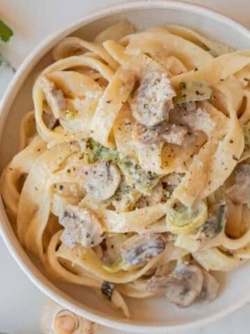 Bowl of creamy leek and mushroom pasta topped with Parmesan cheese and black pepper on a light marble surface.