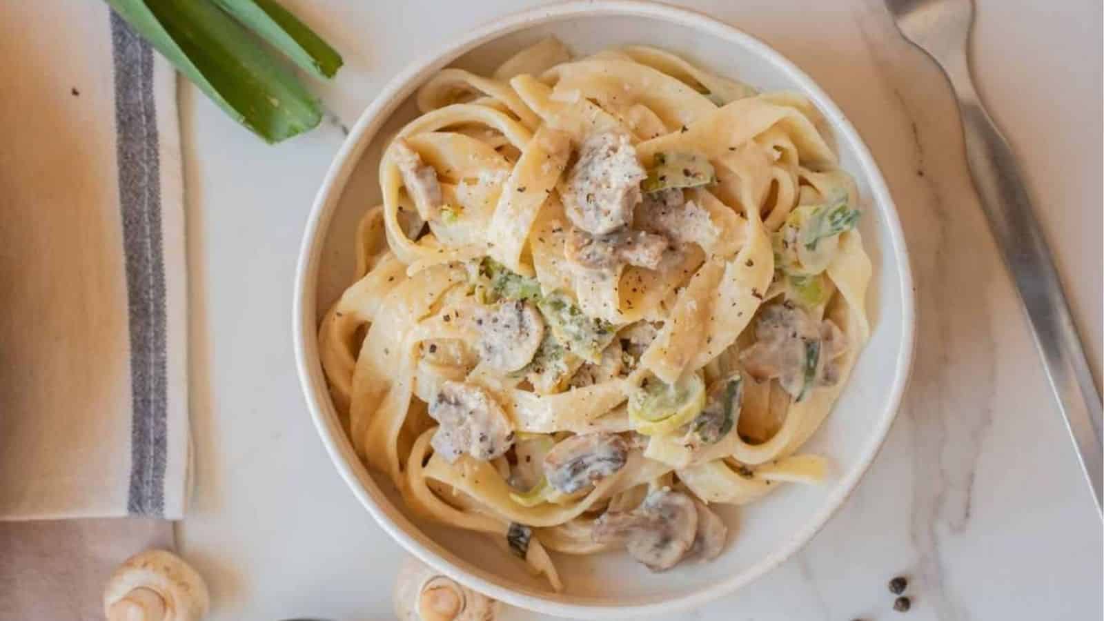 Bowl of creamy leek and mushroom pasta topped with Parmesan cheese and black pepper on a light marble surface.